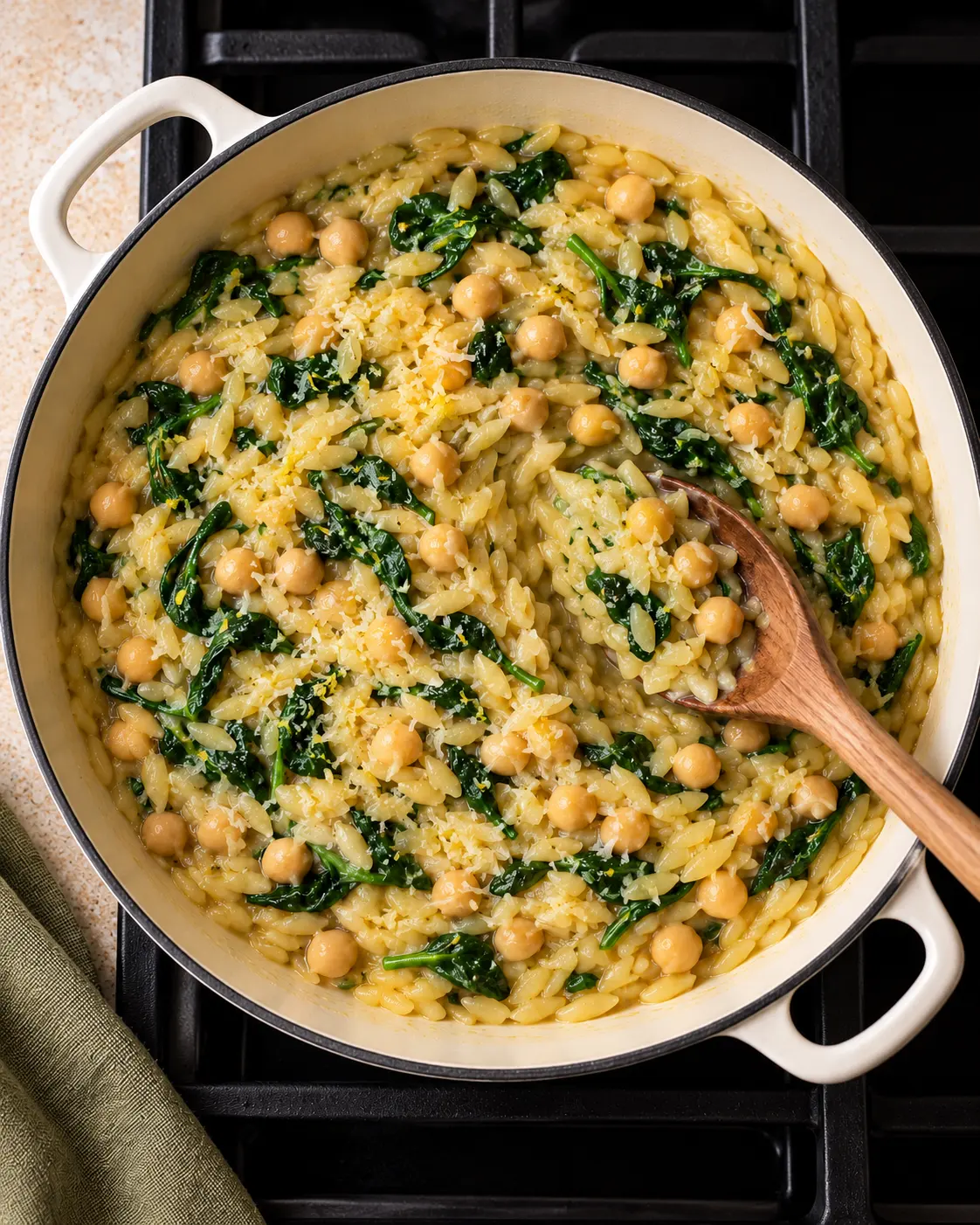 Finished lemon chickpea orzo with wilted spinach and Parmesan in the same cream-colored pan, with a wooden spoon lifting a portion.