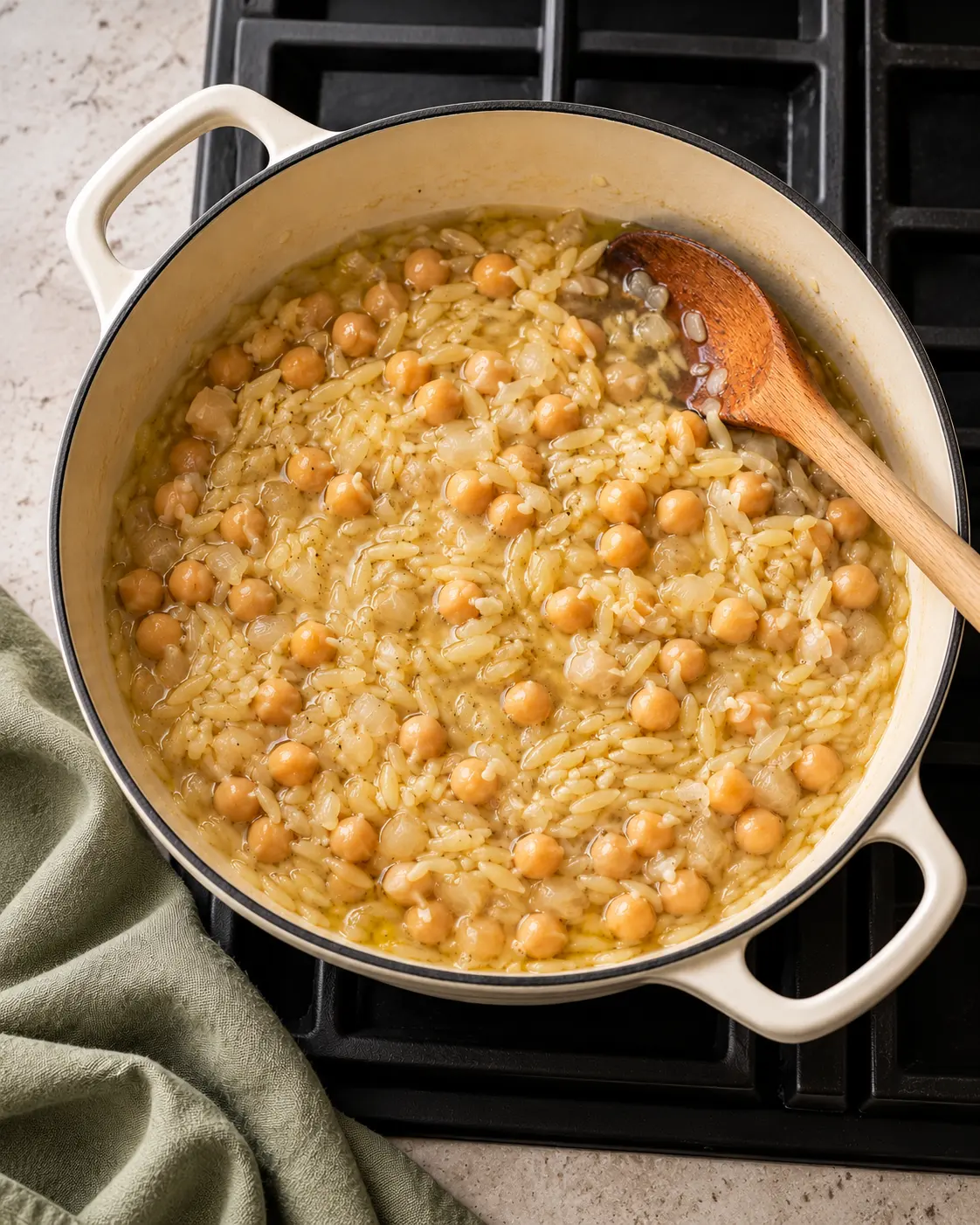 Orzo and chickpeas simmering with diced onion in a cream-colored pan before the spinach is added.